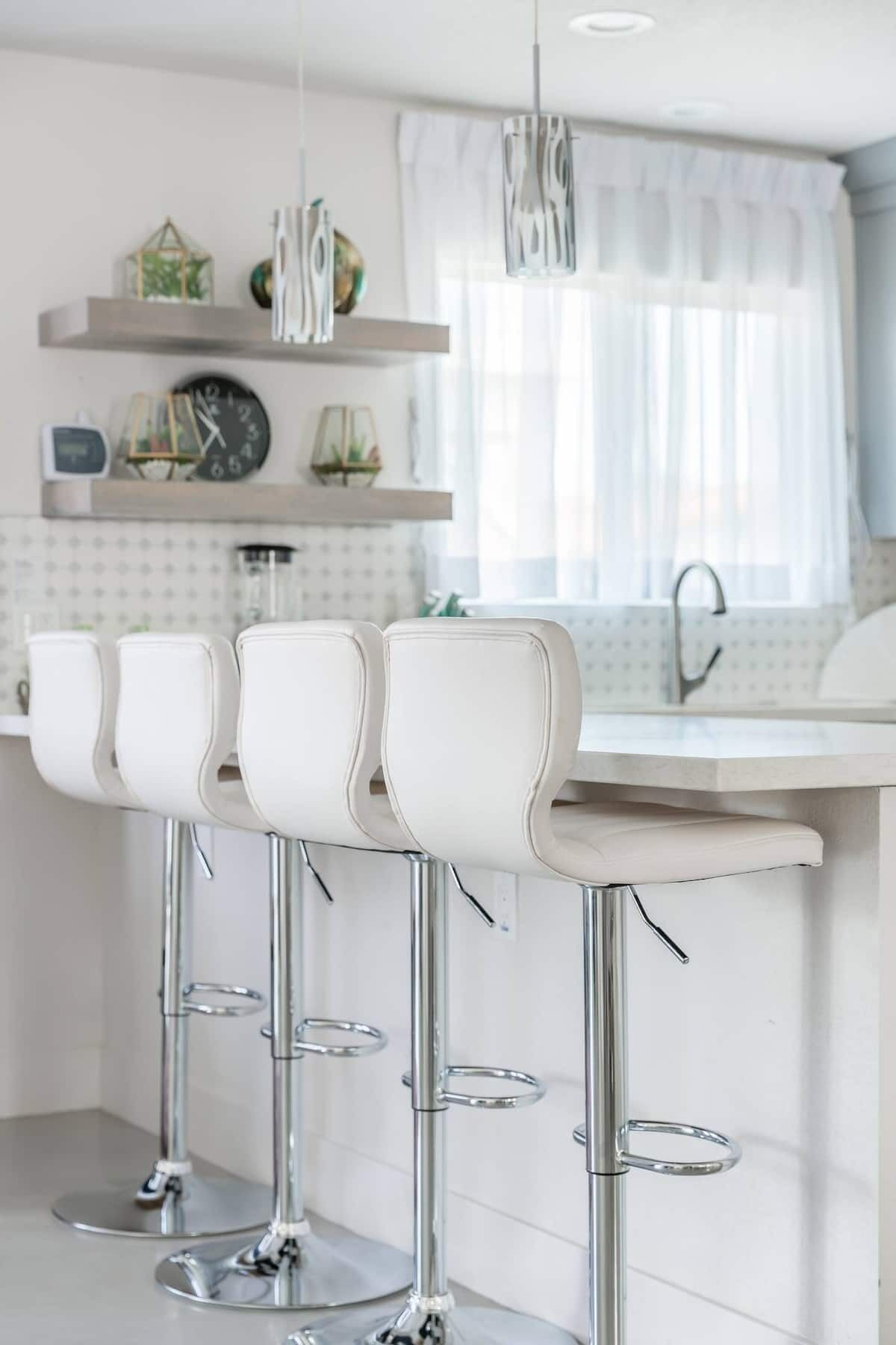 Una cocina moderna con taburetes blancos, un lavamanos de acero inoxidable y luces colgantes.