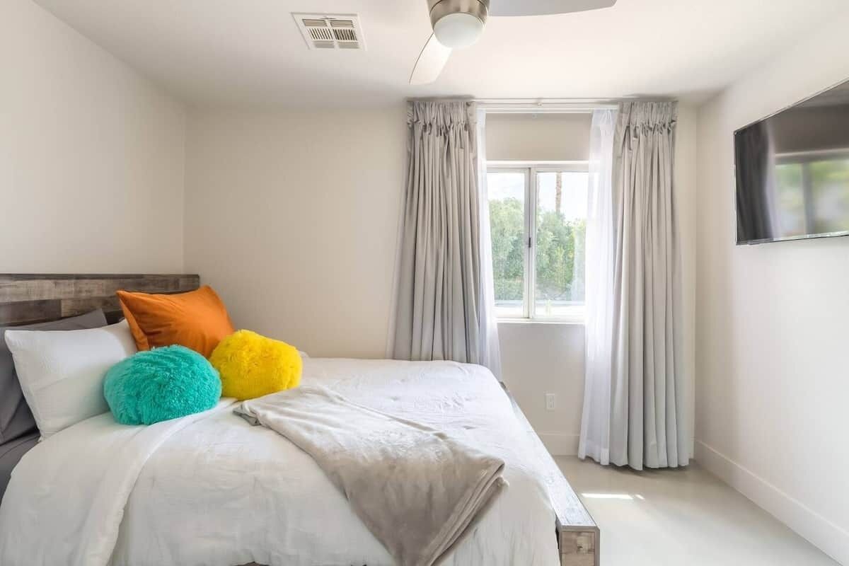 A bedroom with a bed, a ceiling fan, a window with curtains, and a flat-screen TV on the wall.