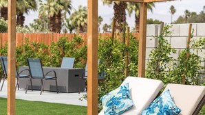 Una pérgola de madera con zona de estar acolchada, un pouf amarillo tejido y un entorno junto a la piscina con sillas y follaje.
