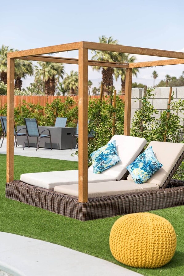 En pergola av trä med en bekväm sittplats med kuddar, en gul stickad pall och en plats vid poolen med stolar och grönska.