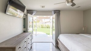 Un dormitorio con cama, televisor montado en la pared, ventilador de techo y vista a un jardín por una puerta abierta.