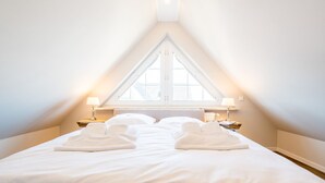 Een net opgemaakt bed met witte lakens en handdoeken, een raam met uitzicht en twee bedlampjes.