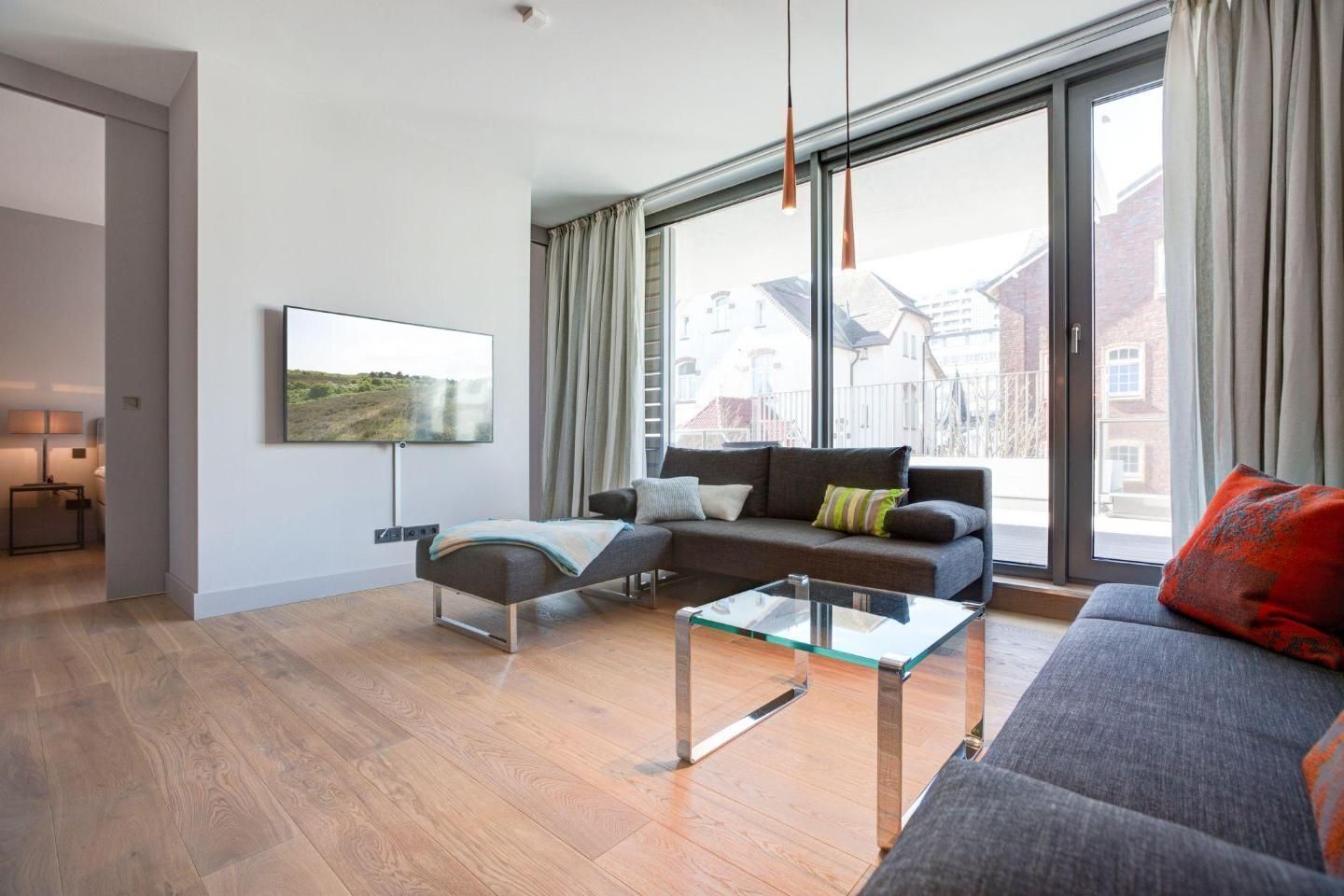 Een moderne woonkamer met een bank, voetenbankje en een glazen salontafel. Een televisie is aan de muur bevestigd en er is een raam met gordijnen.