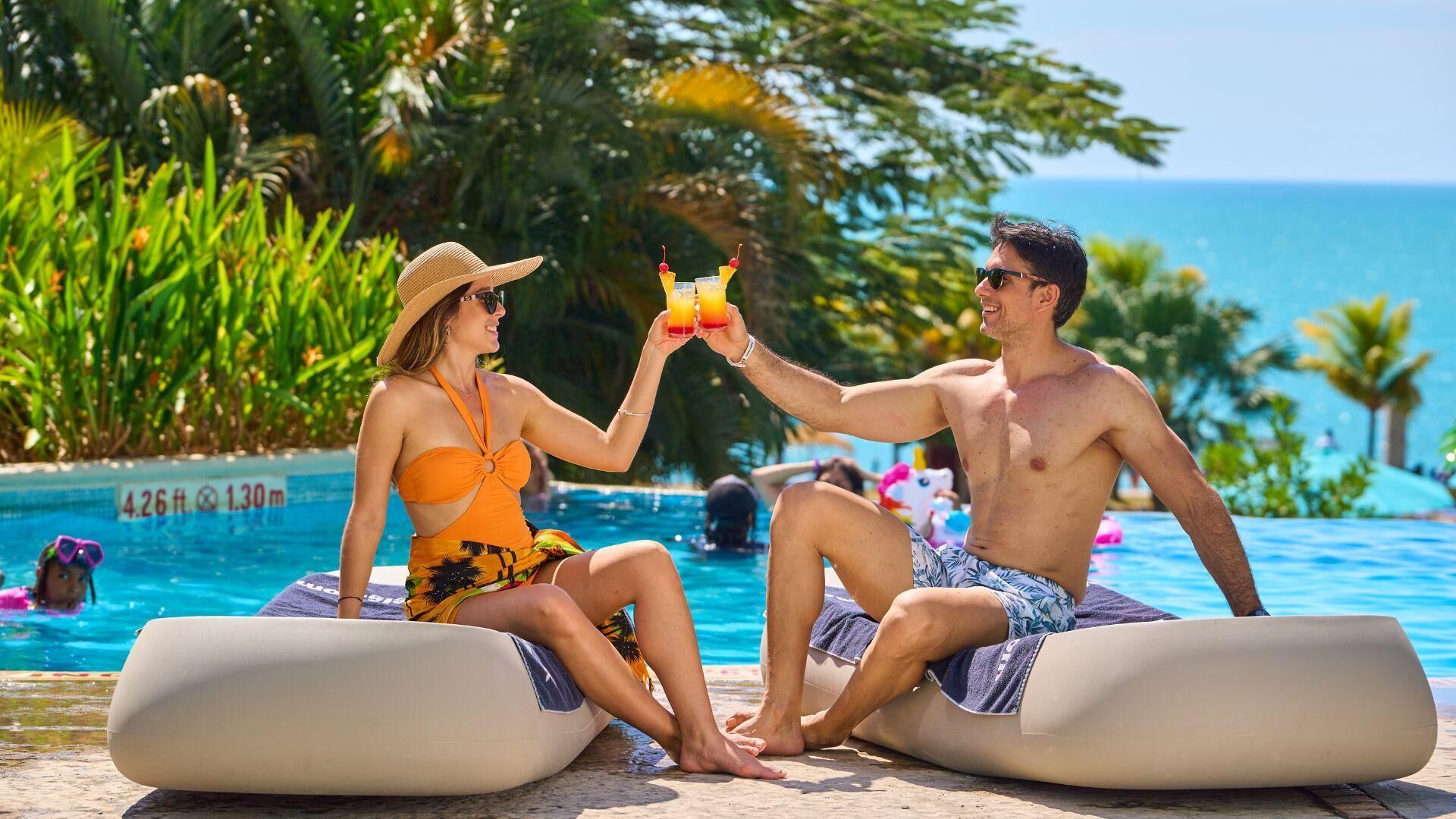 Dos personas sentadas en sillones de descanso junto a una piscina con un paisaje tropical.