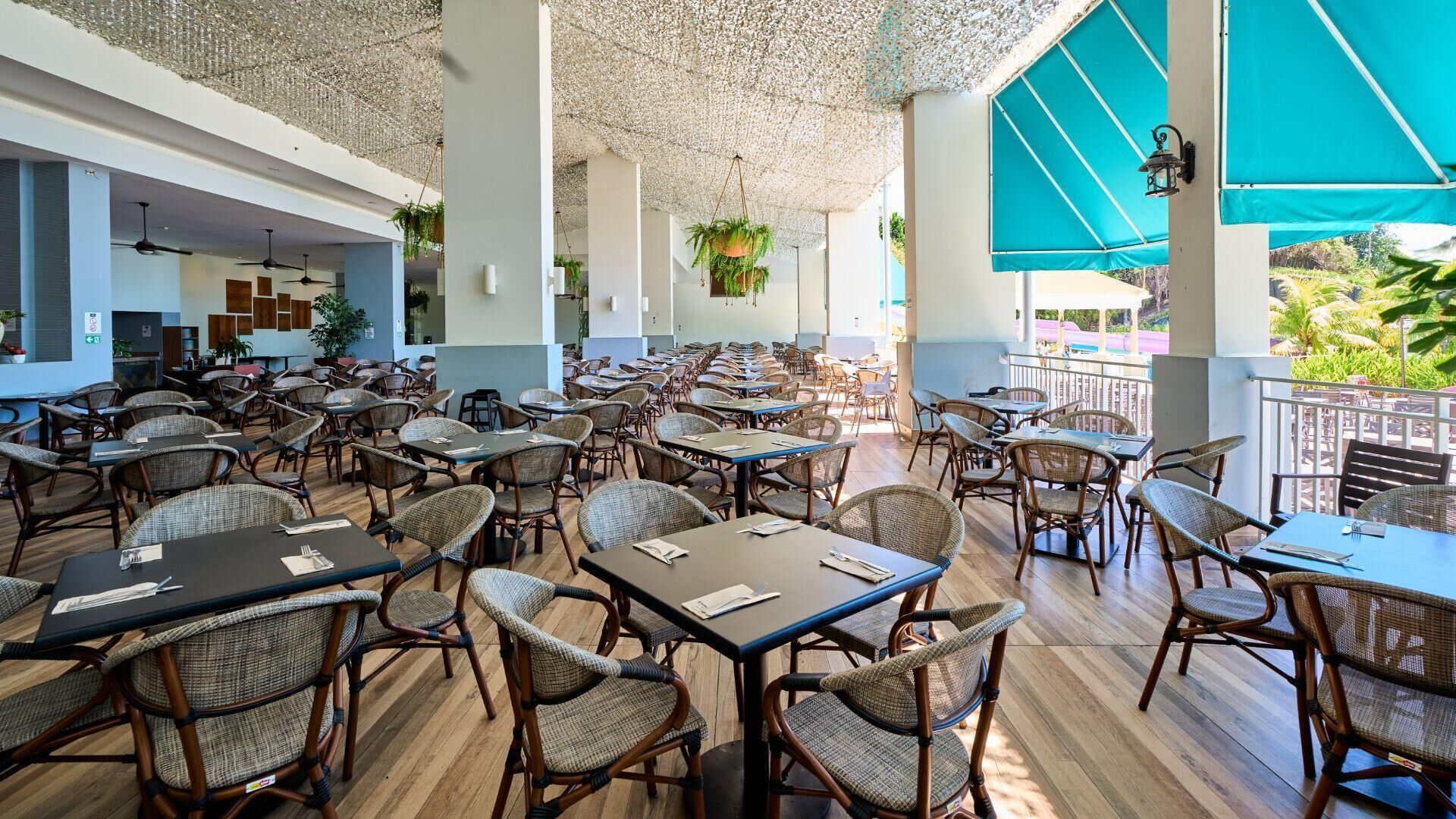 Un amplio espacio exterior para comer con muebles de mimbre y mesas, rodeado por un techo y paredes blancos, con vistas a zonas verdes.