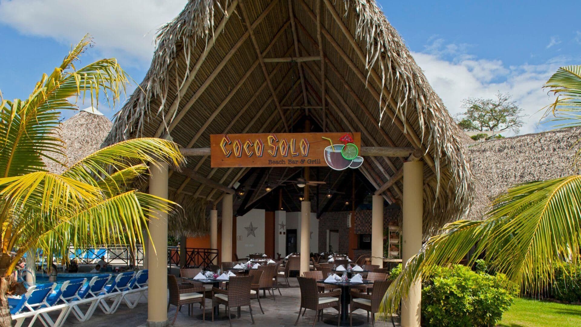 A beach bar and grill named Coco Solo with outdoor seating and a thatched roof.