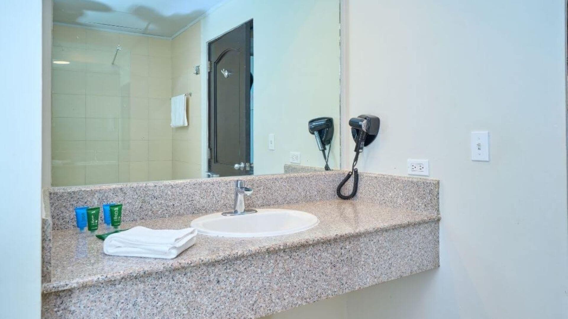 Un baño con encimera de granito, un lavabo, un espejo y dos secadores de pelo fijados a la pared.