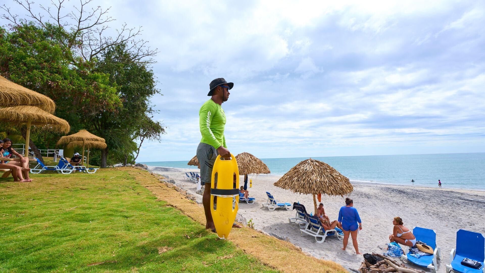 A beach with thatched umbrellas, sun loungers, and a grassy area.