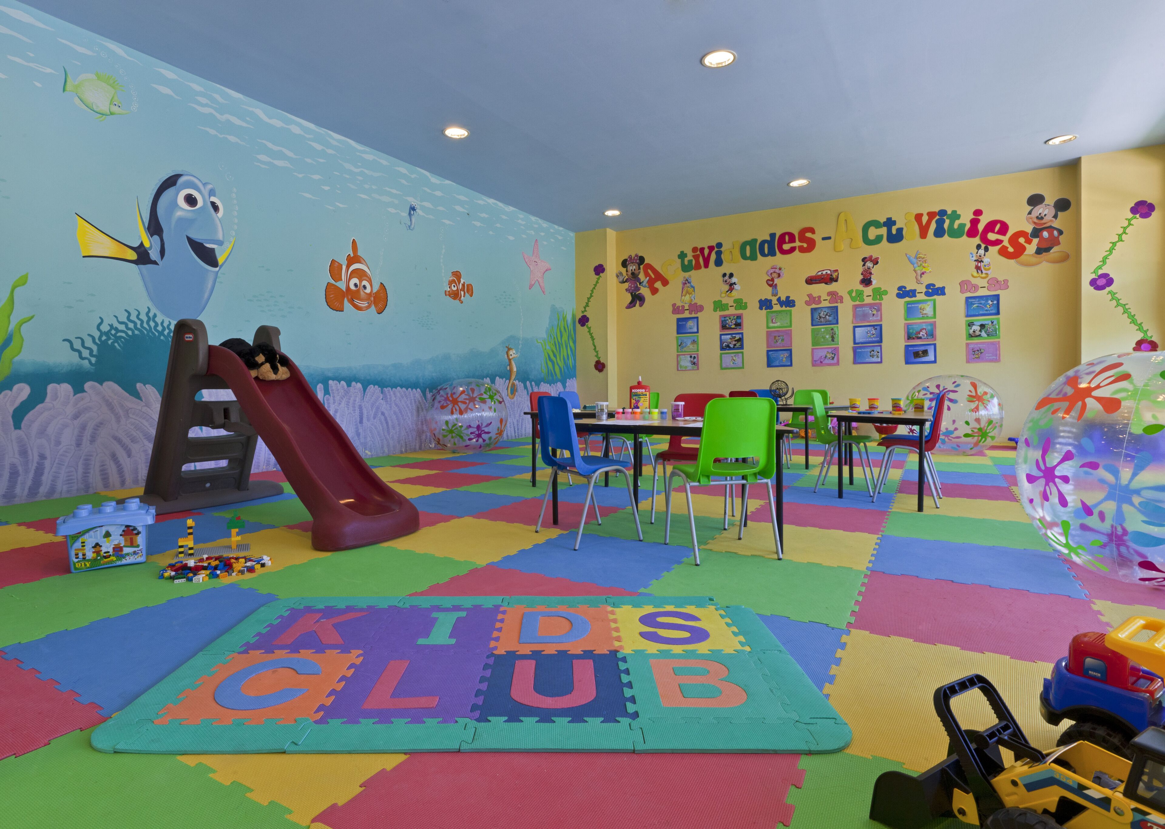 Zona de juegos infantiles con un tobogán, alfombras de juego coloridas y carteles educativos en las paredes.