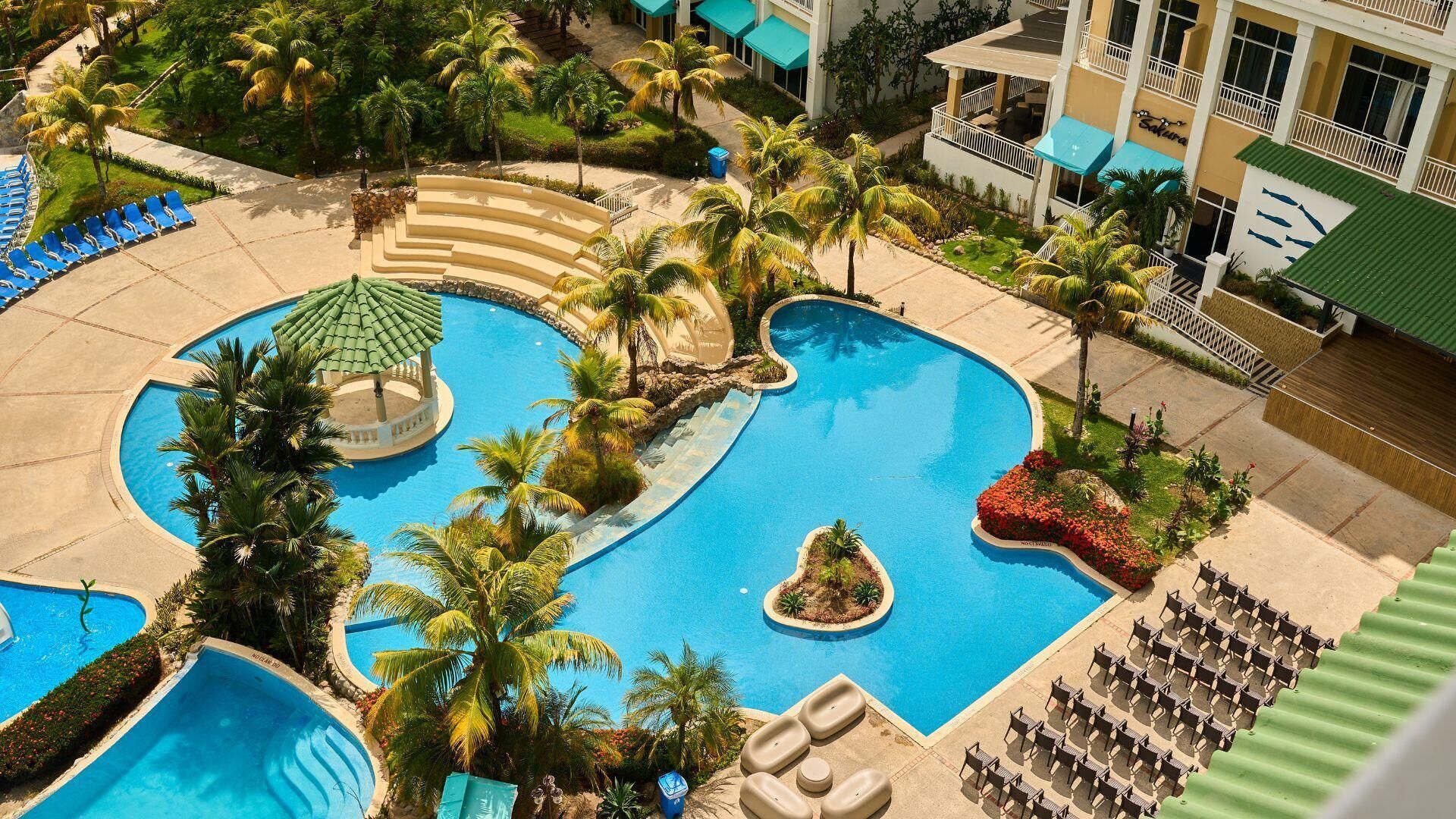 Une grande piscine à plusieurs niveaux, bordée de palmiers et dotée de chaises longues, entourée des bâtiments de l’hôtel.