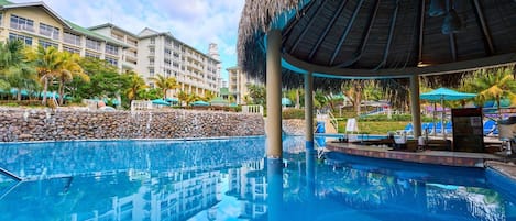 Piscina de un resort con una estructura de techo de paja, una pared de piedra y el reflejo de los edificios del resort en el agua.