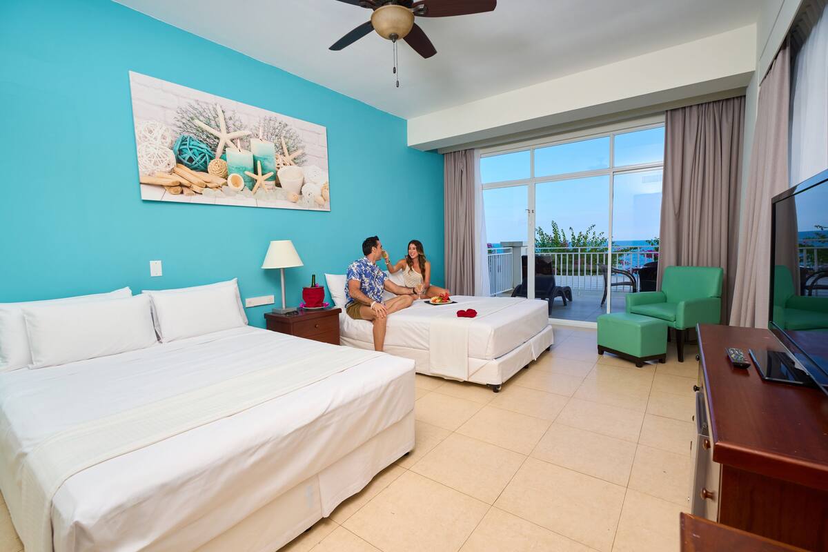 A hotel room with a large bed, a smaller bed, a balcony with a view, a TV, and a painting on the wall.