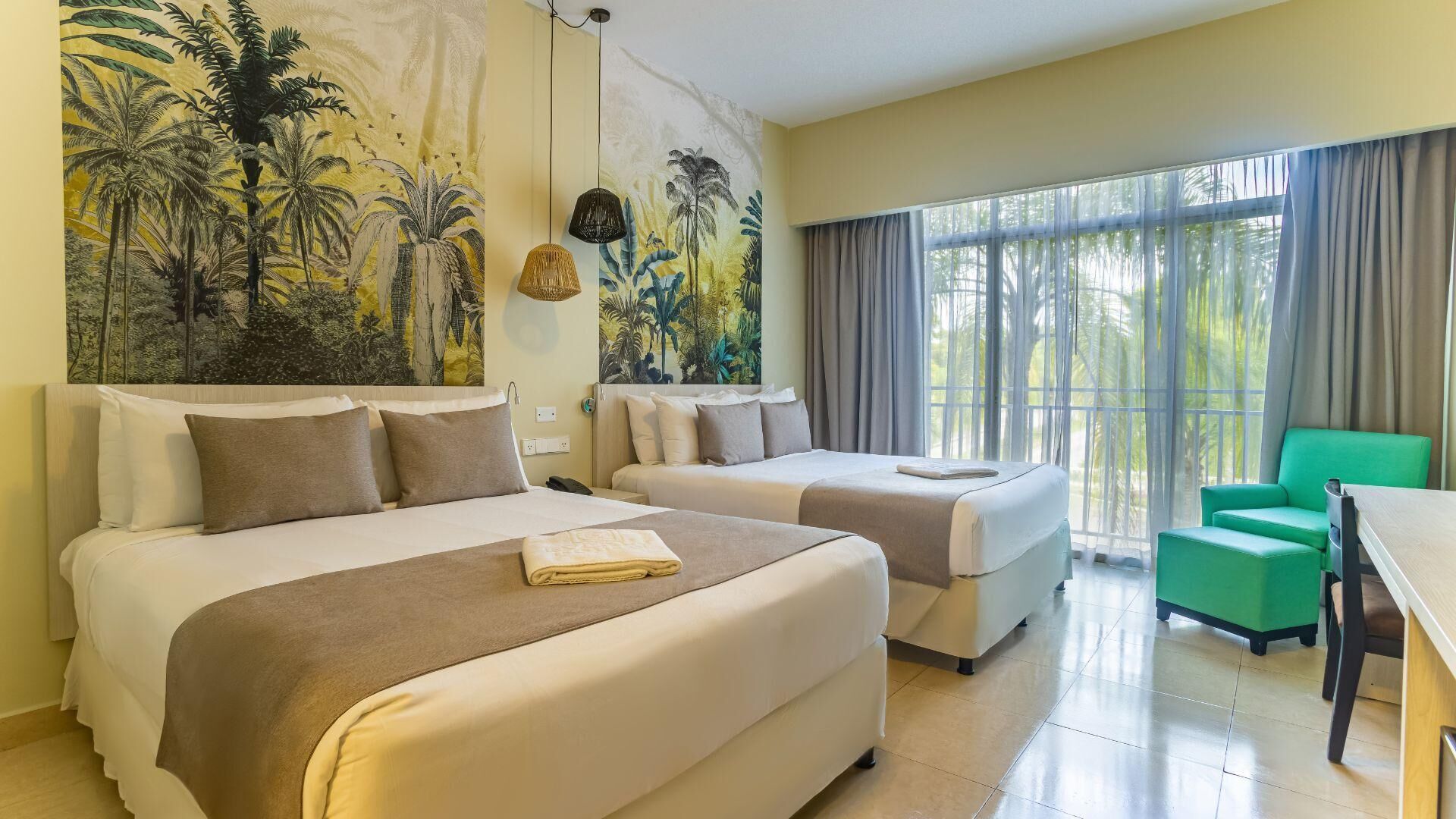 Habitación de hotel con dos camas, una silla verde, una mesa y un gran mural tropical en la pared.