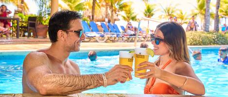 A couple holding drinks at a poolside table with a tropical background.