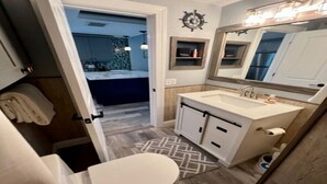 A bathroom with a white sink, vanity, and mirror. There is a towel rack and a small shelf with toiletries. The floor has a geometric patterned rug.