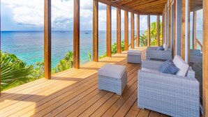 Een houten terras met rieten meubels en een plafondventilator, met uitzicht op een turquoise zee met een zeilboot.