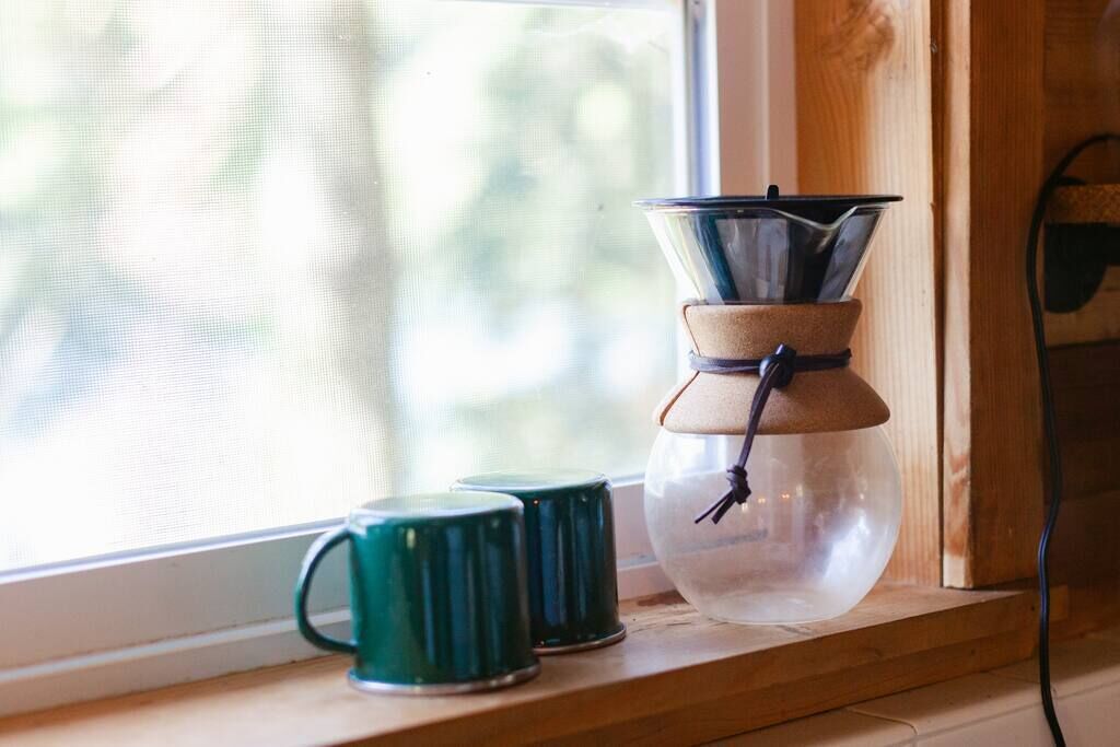Una cafetera de vidrio transparente con base marrón y cable negro, colocada sobre un repisa de madera junto a una ventana.