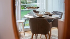 Een eetruimte met een ronde tafel gedekt voor twee personen, een klein bijzettafeltje en uitzicht op een tuin door een groot raam.
