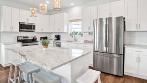 Una cocina moderna con armarios blancos, electrodomésticos de acero inoxidable, una isla de mármol e iluminación colgante.