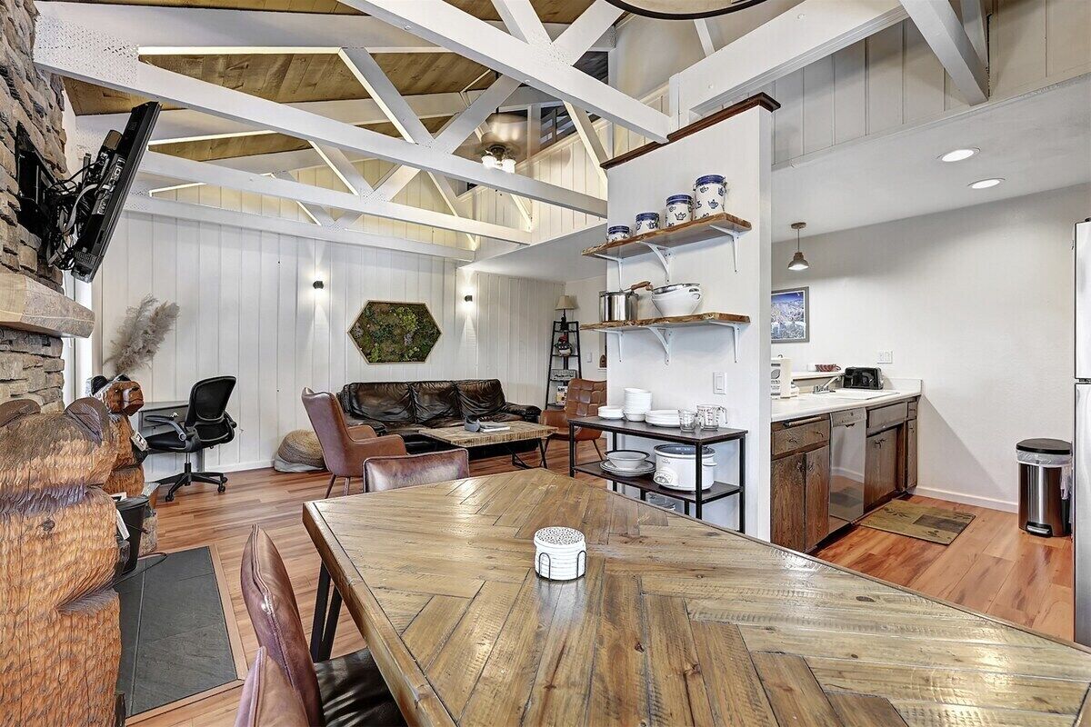 A spacious living area with a wooden dining table, a stone fireplace, and a kitchen with white cabinetry and a hanging pot rack.