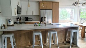 Een moderne keuken met witte kasten, een groot eiland met krukken, roestvrijstalen apparatuur en uitzicht op de omgeving.