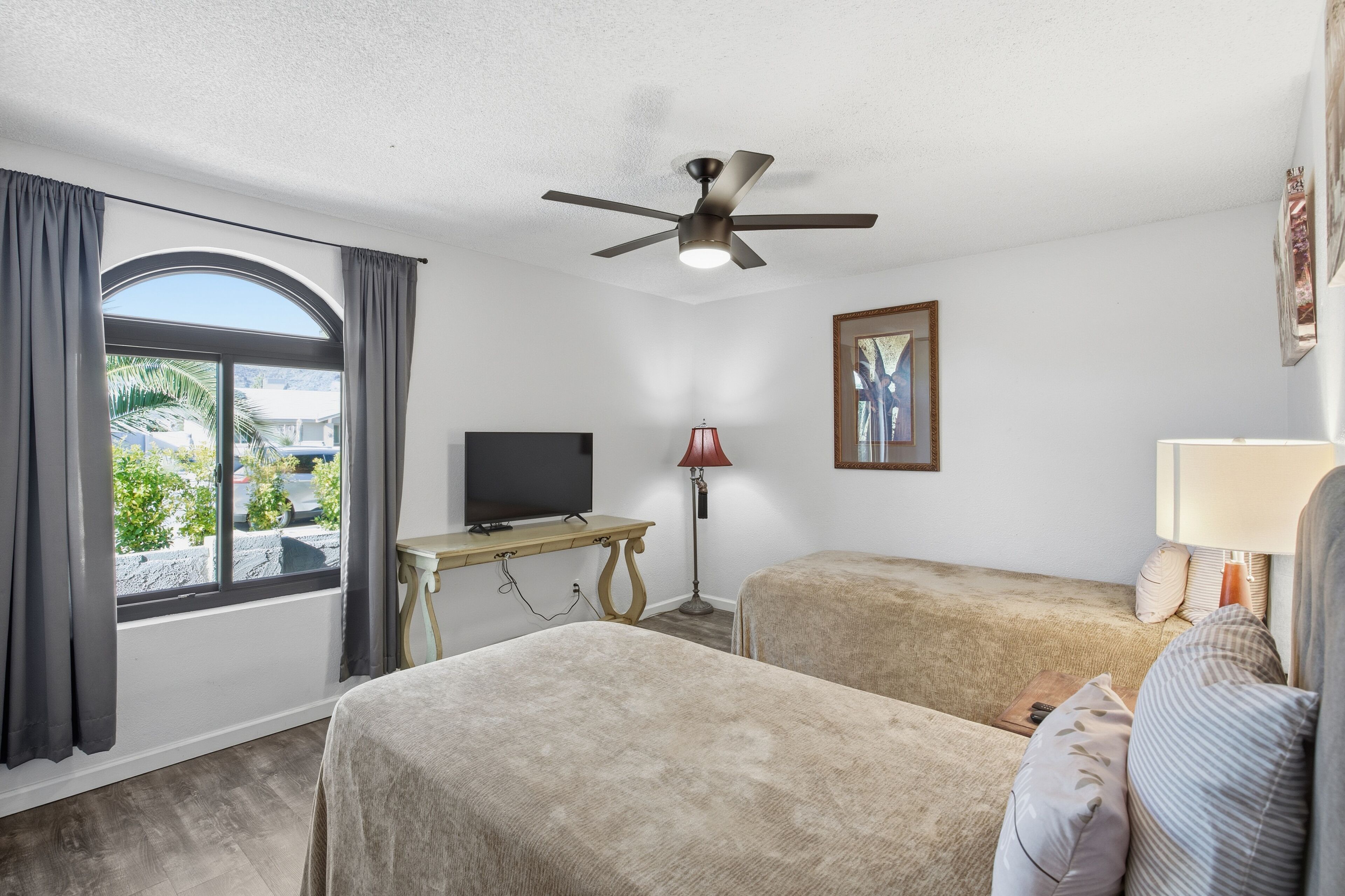 A hotel room with two beds, a ceiling fan, a TV on a wooden table, a lamp, and a framed picture on the wall.