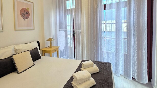 A neatly made bed with white and black bedding, a bedside table with a lamp, and a window with sheer curtains. - Apt Porto Ribeira Rio Douro - 6 Guests  (Porto)