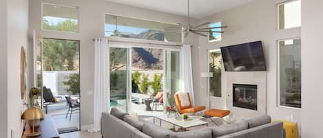 A spacious living room with a gray sectional sofa, a coffee table, and a fireplace. There is a large window offering a view of a pool and mountains.