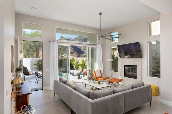 A spacious living room with a gray sectional sofa, a coffee table, and a fireplace. There is a large window offering a view of a pool and mountains.