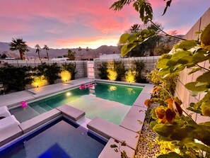 A pool area with two pools, surrounded by plants and palm trees, and a sunset in the background.