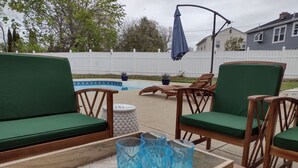 A poolside area with wooden chairs and a green sofa, a wooden table with blue glasses, and a white fence in the background.