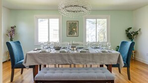 A dining room with a table set for six, two blue chairs, a bench, a chandelier, and a framed picture on the wall.