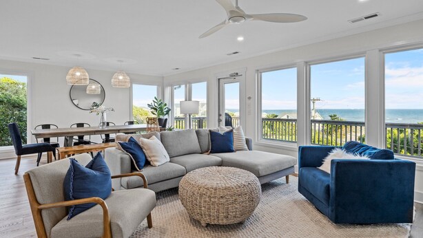 A spacious living room with a large sectional sofa, a round ottoman, and a blue armchair. There is a dining area with a table and chairs, and a balcony with a view of the ocean. - OCEAN VIEWS | 2nd Row | Heart of EI | NEWLY RENOVATED!! (Emerald Isle)