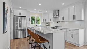 A modern kitchen with white cabinetry, stainless steel appliances, and a large island with bar stools. - OCEAN VIEWS | 2nd Row | Heart of EI | NEWLY RENOVATED!! (Emerald Isle)