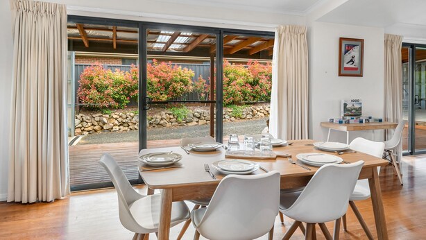 Een eethoek met een tafel voor vier personen, grote ramen en uitzicht op een tuin met herfstkleuren.