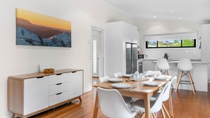 Een moderne keuken met witte kasten, een eethoek met een houten tafel en witte stoelen, en een groot schilderij van een berglandschap aan de muur.