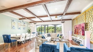 Un salon spacieux avec un plafond en bois, un grand ensemble de canapés, un coin repas avec des chaises blanches et une vue sur la verdure à l’extérieur.