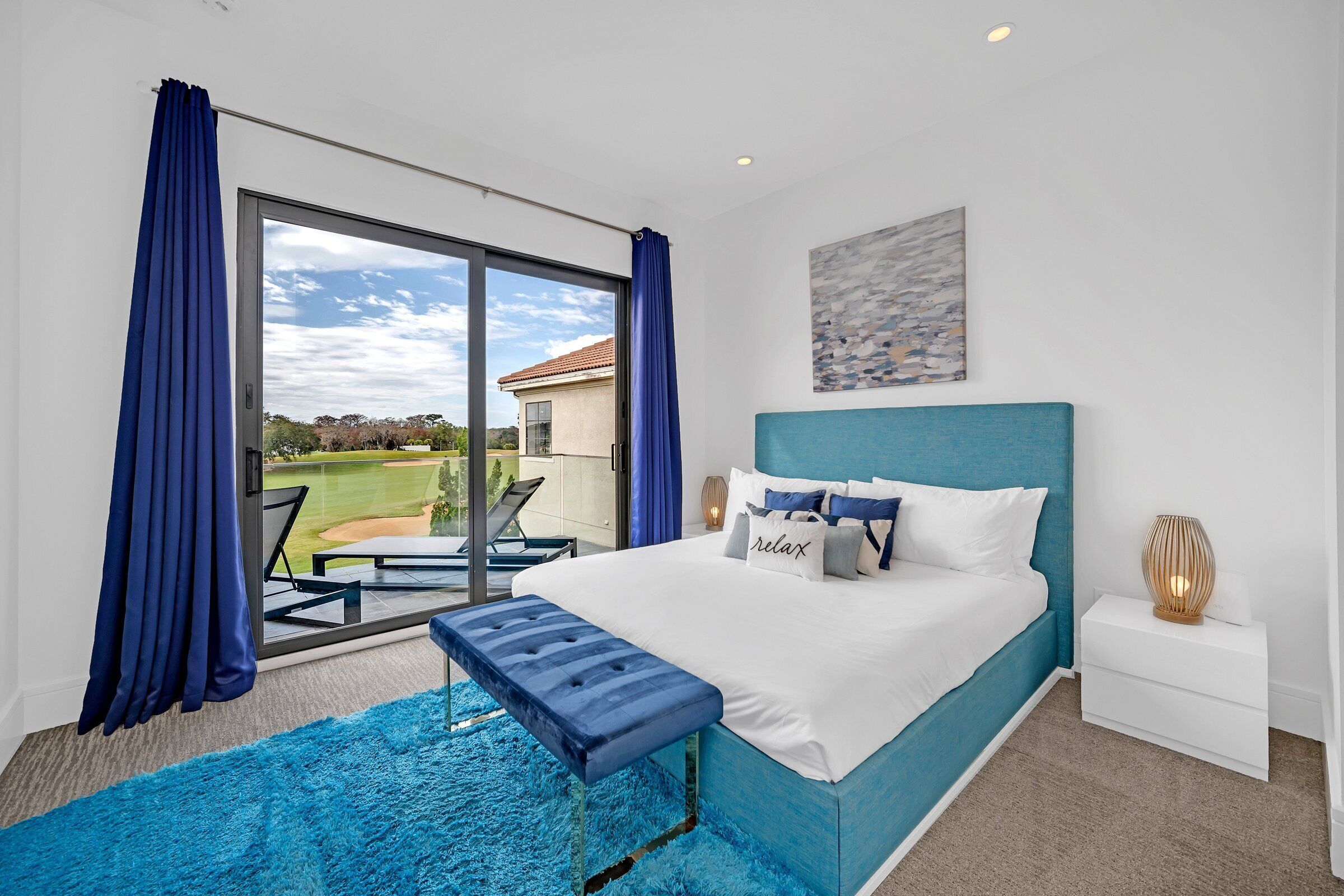 A bedroom with a large window, a bed with a blue headboard, a bench, and a nightstand with a lamp.