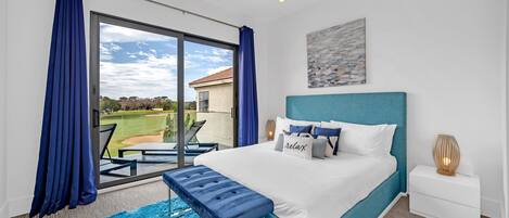 A bedroom with a large window, a bed with a blue headboard, a bench, and a nightstand with a lamp.