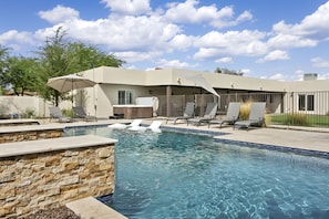 A swimming pool with lounge chairs and umbrellas, surrounded by a stone wall and a building with large windows.