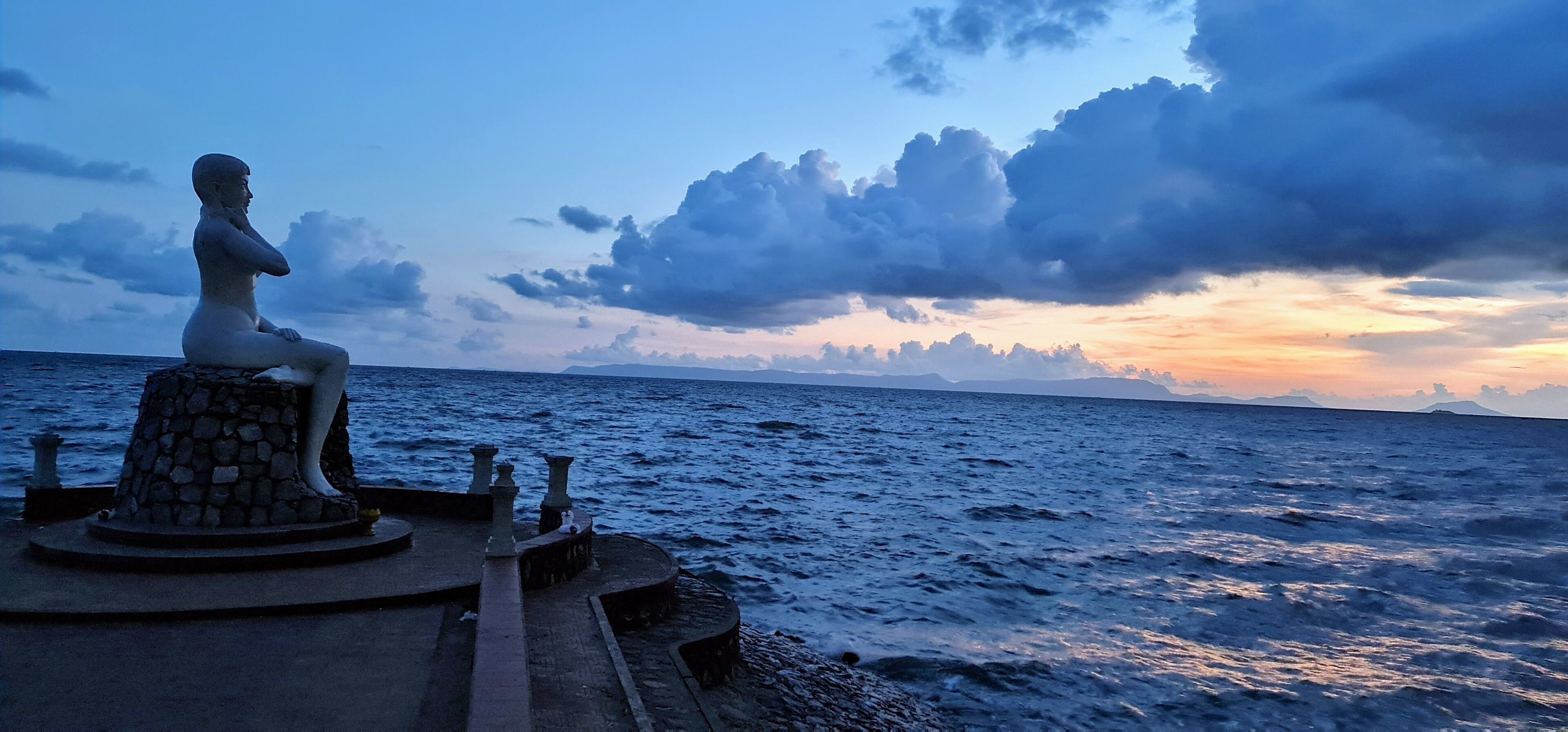 A statue on a stone platform by the sea with a view of the ocean and a sunset sky.