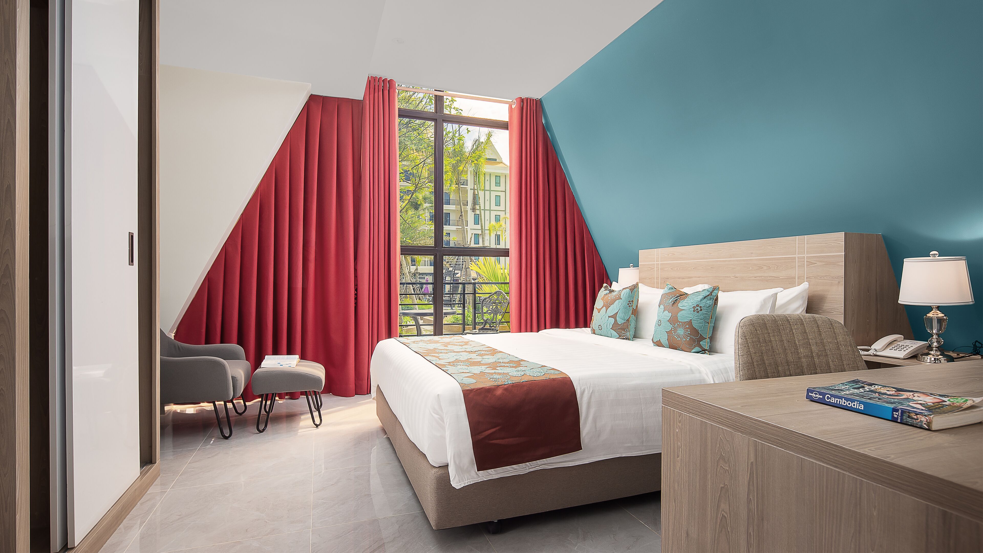 Ein Hotelzimmer mit einem großen Bett, einem Schreibtisch mit Lampe, einem Sessel und Blick auf Gebäude durch ein Fenster mit roten Vorhängen.