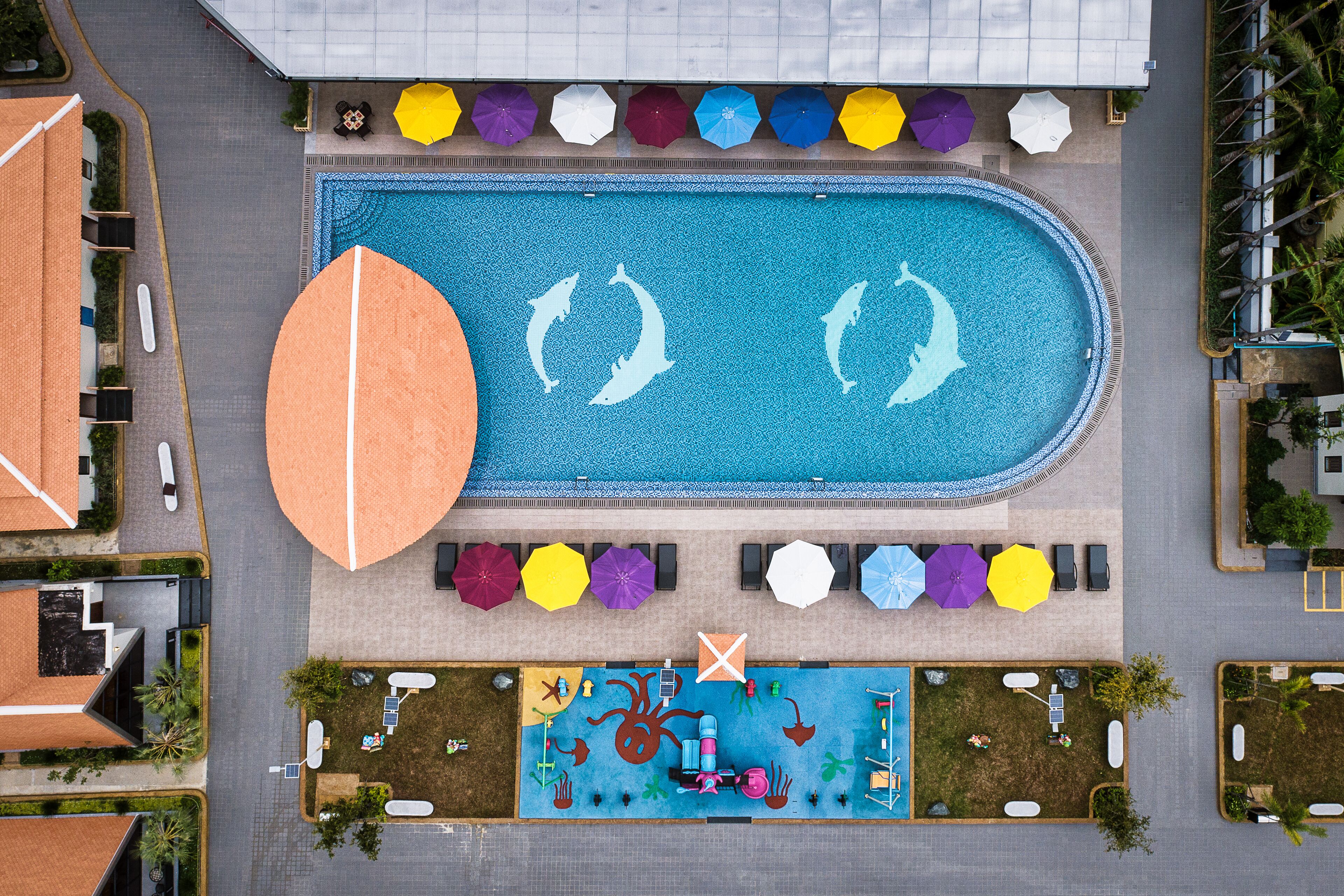 An aerial view of a hotel with a swimming pool, sun loungers, umbrellas, and a children's play area.