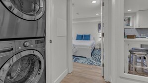 Un espace buanderie avec des machines à laver superposées, une chambre avec un lit et des oreillers bleus, et un coin cuisine avec un tabouret.