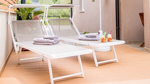 Un transat avec un parasol blanc, une petite table avec une serviette et un pot de fleurs suspendu.