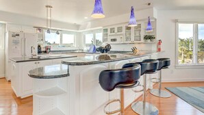 A modern kitchen with white cabinetry, a large island with bar stools, pendant lighting, and a view of the outdoors. - Shore Elegance | Oceanfront | Private Pool (Virginia Beach)