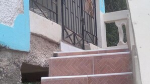 A staircase leading to a door with a metal gate, part of a building with a textured wall and a window with a decorative frame.