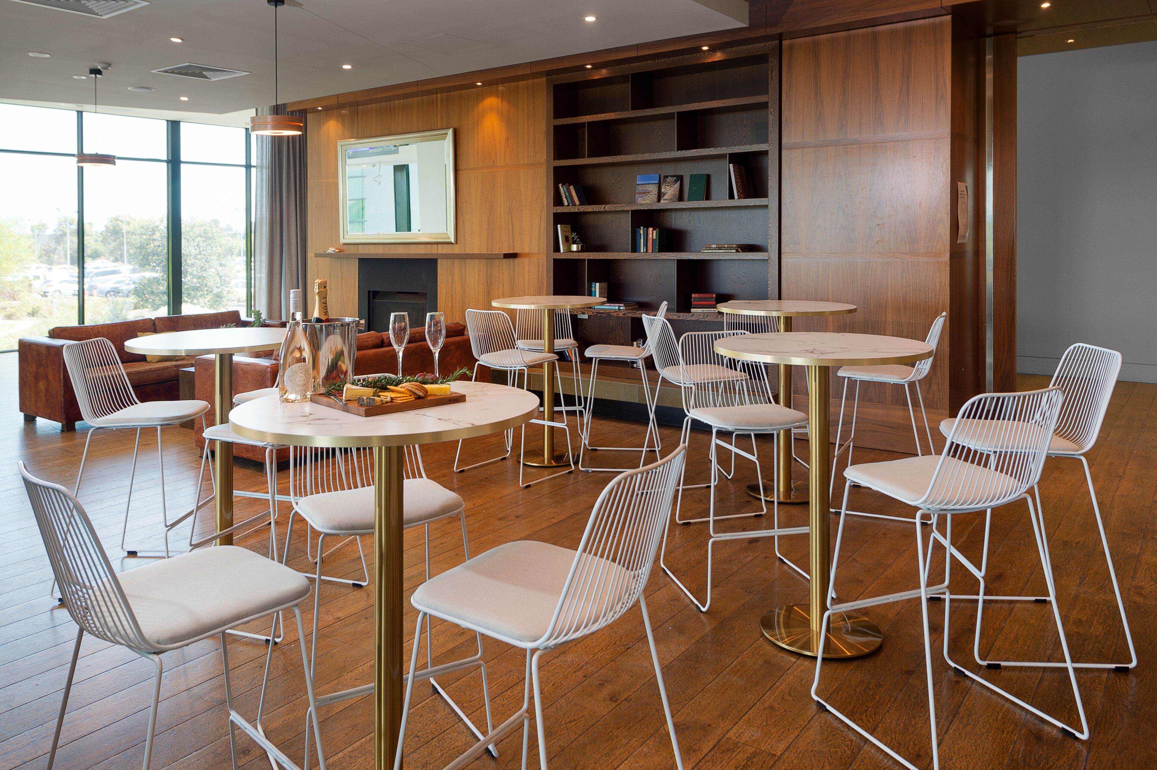 Un comedor moderno con mesas redondas y sillas de alambre blanco, pisos de madera y una estantería con libros y objetos decorativos.