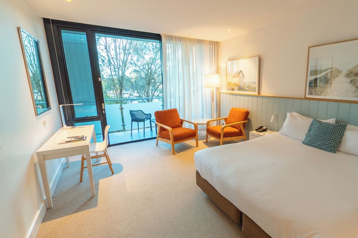 Ein Hotelzimmer mit einem großen Bett, zwei Sesseln, einem kleinen Tisch und einem Schreibtisch. Ein Fenster bietet Blick auf Bäume und eine Tür führt zu einem Balkon.