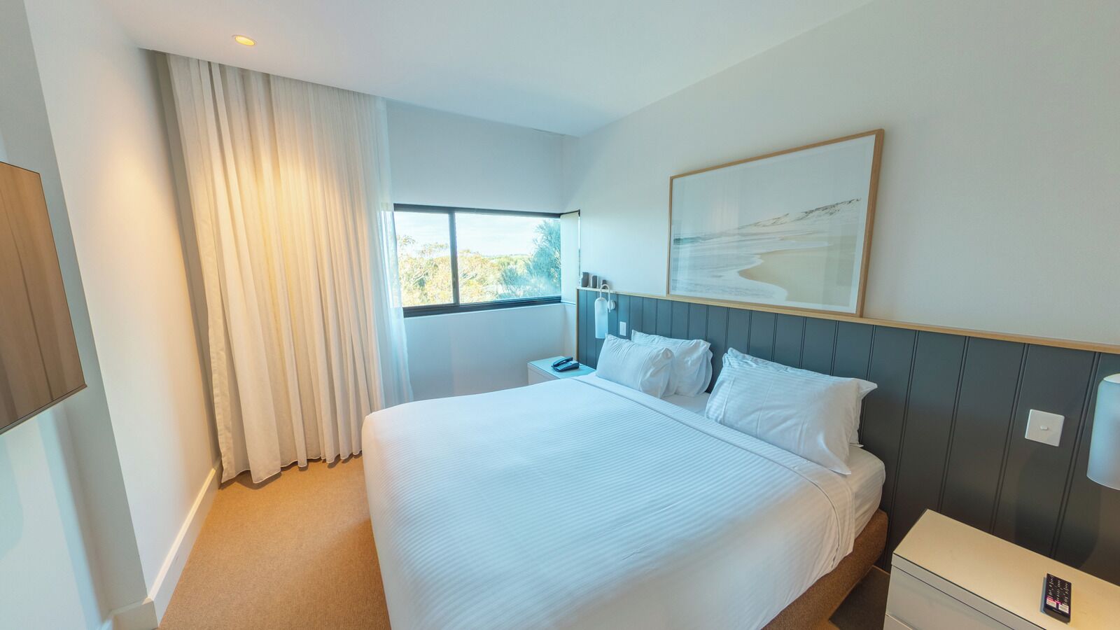A hotel room with a large bed, a bedside table, a nightstand, a window with curtains, and a framed picture on the wall.